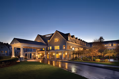 The Village at Duxbury senior living facility building at dusk with warm interior lights glowing through the windows, a covered entrance driveway, well-maintained landscaping, and a clear evening sky.