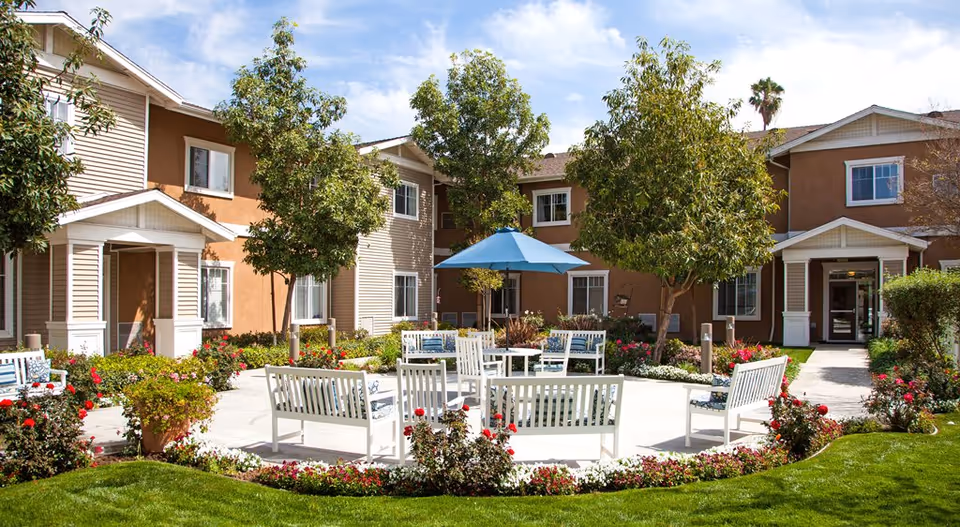 Sunny courtyard with white benches arranged around a patio, flower beds, a blue umbrella, trees, and the two-story building facade behind.