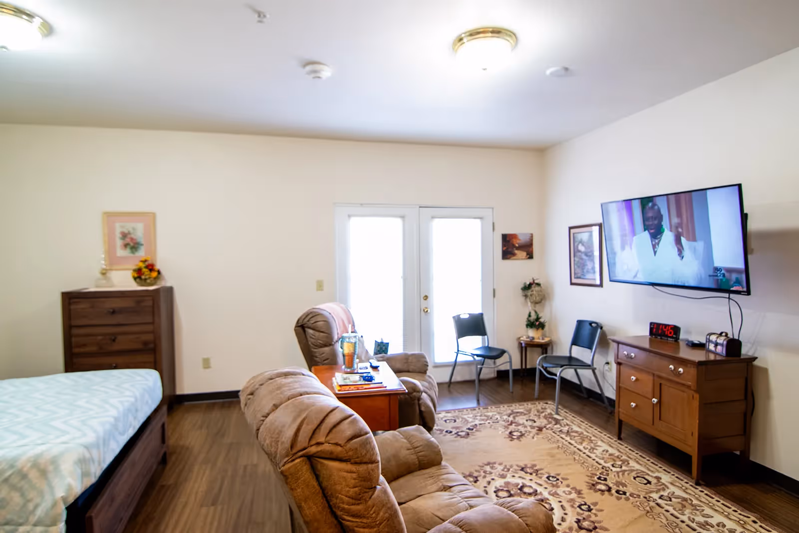 A cozy living area in a senior living facility featuring a brown recliner chair, a wooden side table with a drink and books, a flat-screen TV mounted on the wall, two black chairs, a wooden dresser, and a bed partially visible on the left. The room has light-colored walls, wooden flooring, a patterned area rug, and double glass doors letting in natural light.