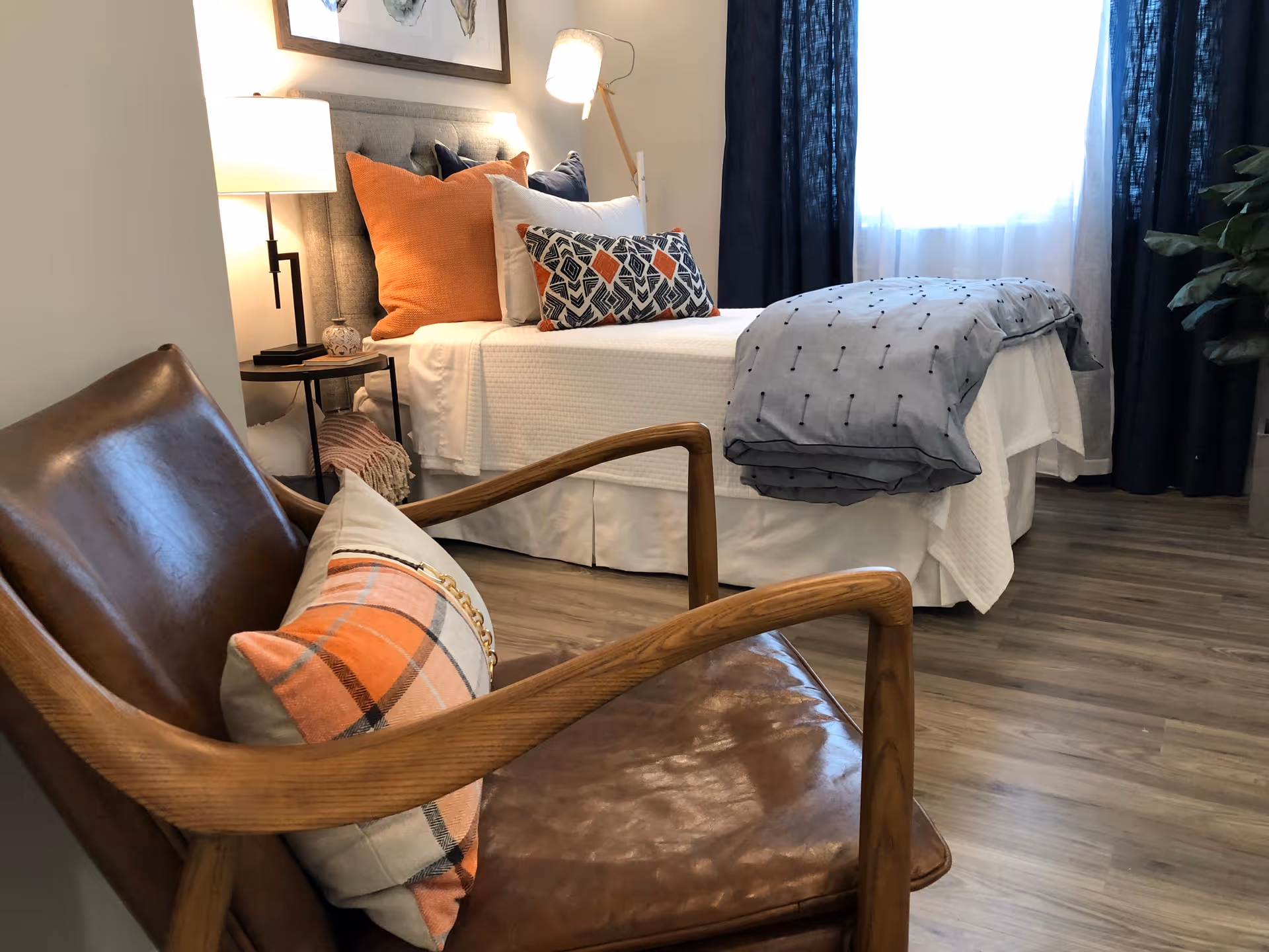 A cozy bedroom featuring a neatly made bed with white bedding, orange and patterned pillows, and a gray blanket. Next to the bed is a small round side table with a lamp and decorative items. In the foreground, there is a brown leather chair with wooden armrests and an orange plaid pillow. The room has wood flooring, a window with sheer and dark curtains, and a potted plant in the corner.