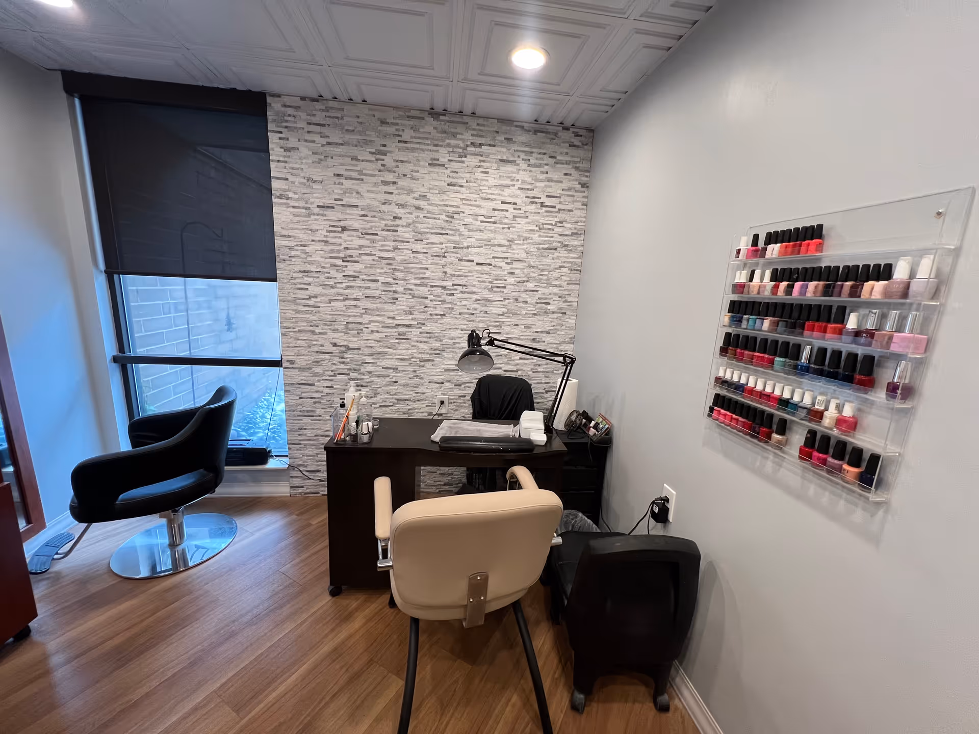 A small nail salon area with a desk and two chairs, one black and one beige. On the desk are nail care items and a desk lamp. A wall-mounted rack displays multiple bottles of nail polish in various colors. The room has wood flooring, a window with a black roller shade, and a textured gray and white accent wall behind the desk.