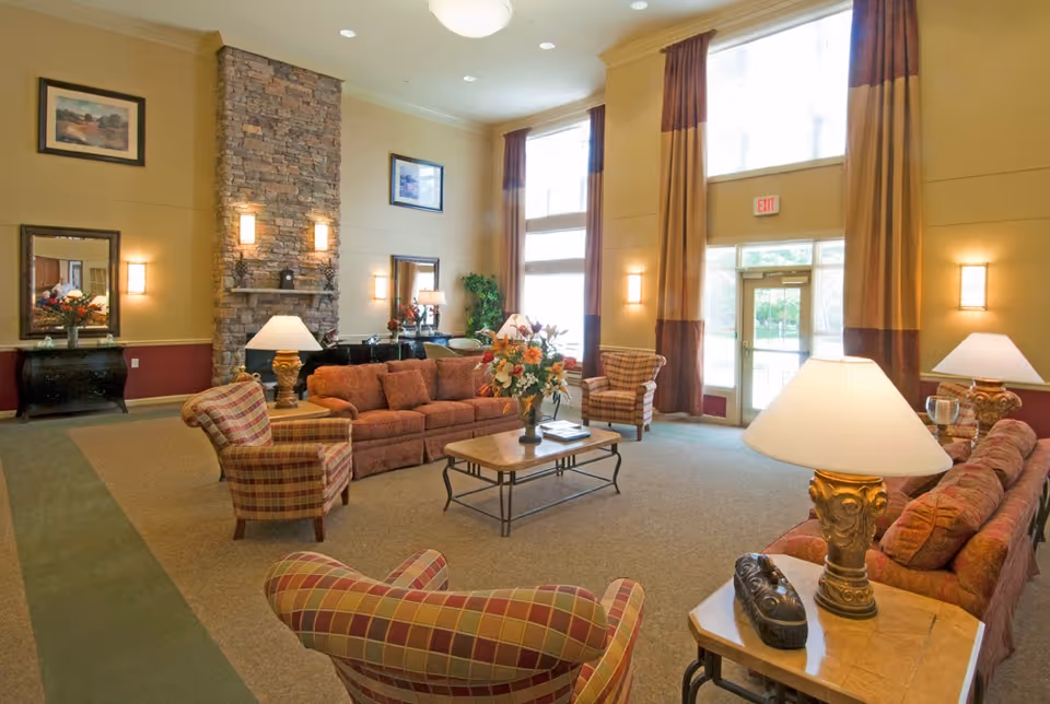 A spacious senior living facility common area with high ceilings, large windows with tall curtains, and a stone fireplace. The room is furnished with multiple cushioned sofas and armchairs in warm tones, a coffee table with a floral arrangement, side tables with lamps, and framed artwork on the walls. The entrance door is visible with an exit sign above it.
