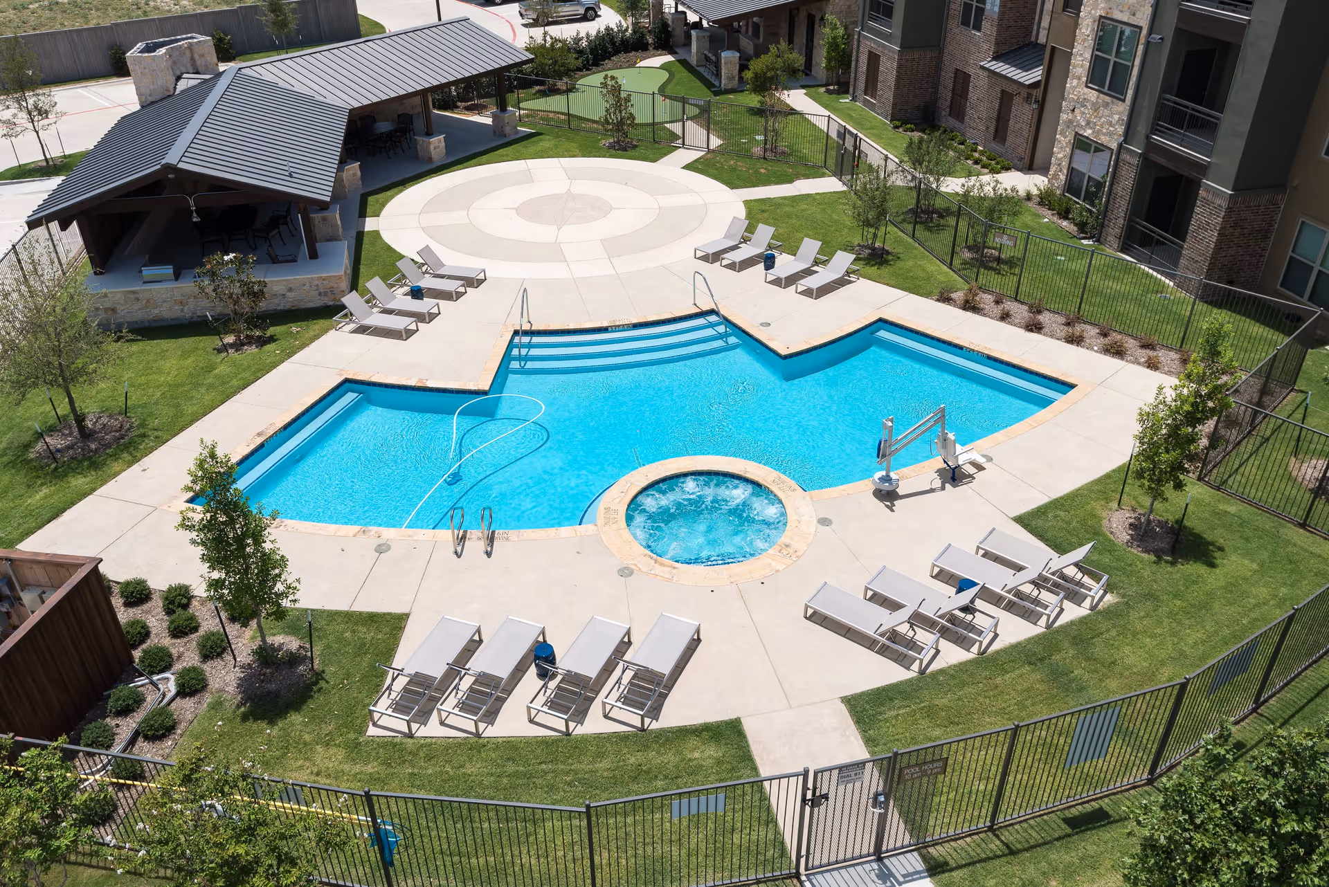Aerial view of an outdoor swimming pool area at a senior living facility. The pool has a unique geometric shape with a circular hot tub attached. Surrounding the pool are several lounge chairs arranged on a concrete deck. There is a covered pavilion with seating and a stone fireplace to the left. The area is fenced and landscaped with grass, trees, and shrubs. Adjacent to the pool area are multi-story residential buildings.