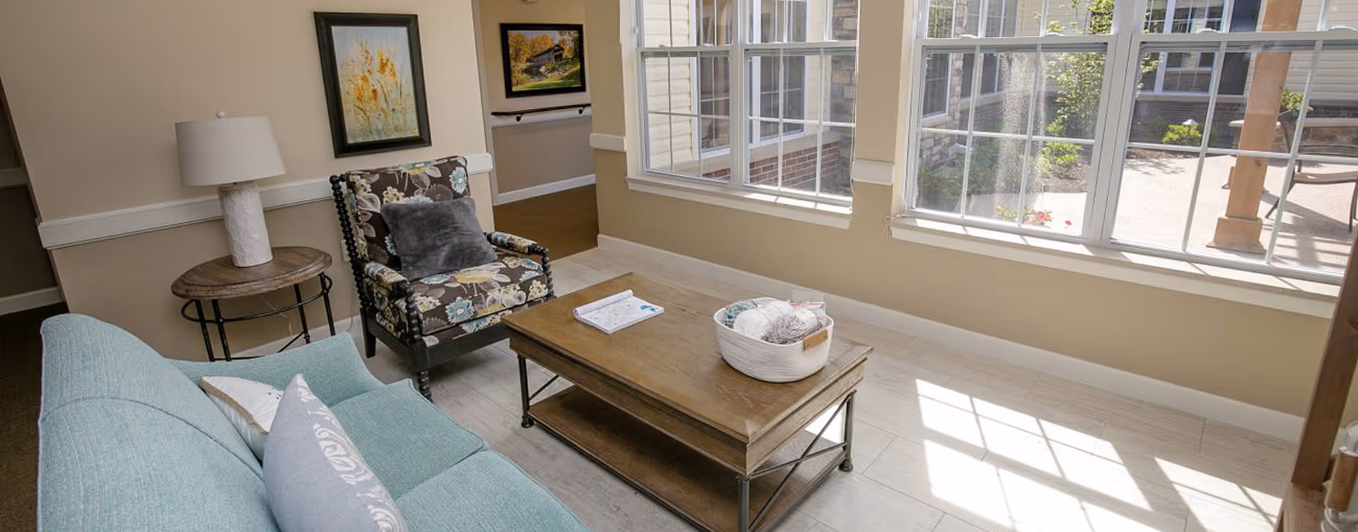 A cozy living room area with a light blue sofa adorned with pillows, a floral patterned armchair with a gray cushion, a wooden coffee table with a white basket containing yarn balls, and a round side table with a white lamp. Large windows let in natural light and offer a view of an outdoor patio area.