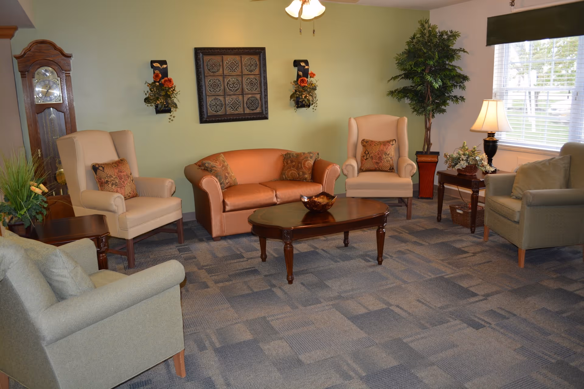 A cozy communal living room with sofas and armchairs arranged around a wooden coffee table, decorative wall art, plants, and a window with a lamp.