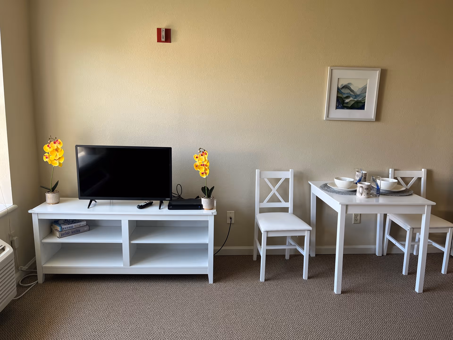 A small living area with a white TV stand holding a flat-screen television, two yellow orchid plants, and some books. Next to it is a white dining table set with two chairs, placemats, bowls, and mugs. A framed abstract painting hangs on the beige wall above the dining table.