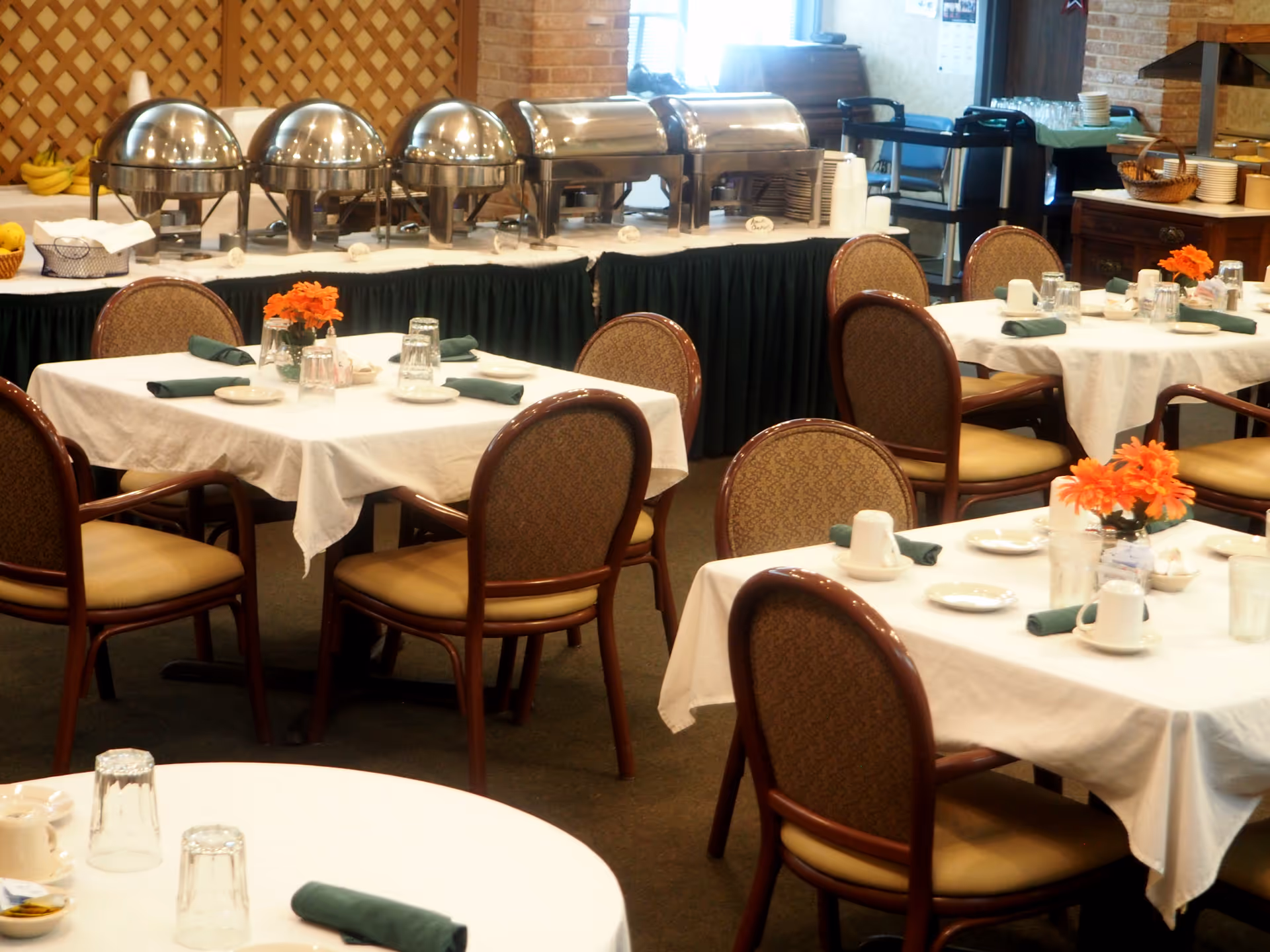 Dining room with several tables covered in white tablecloths, each set with plates, cups, glasses, and green napkins. Orange flowers in small vases decorate the tables. In the background, there is a buffet table with covered chafing dishes and stacks of plates.