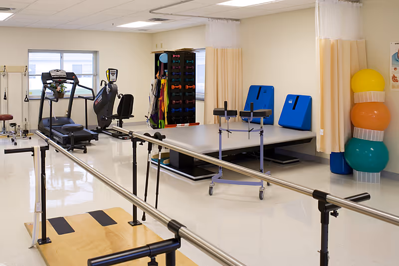 Physical therapy room with parallel bars, exercise equipment, a treatment table, and stacked therapy balls.