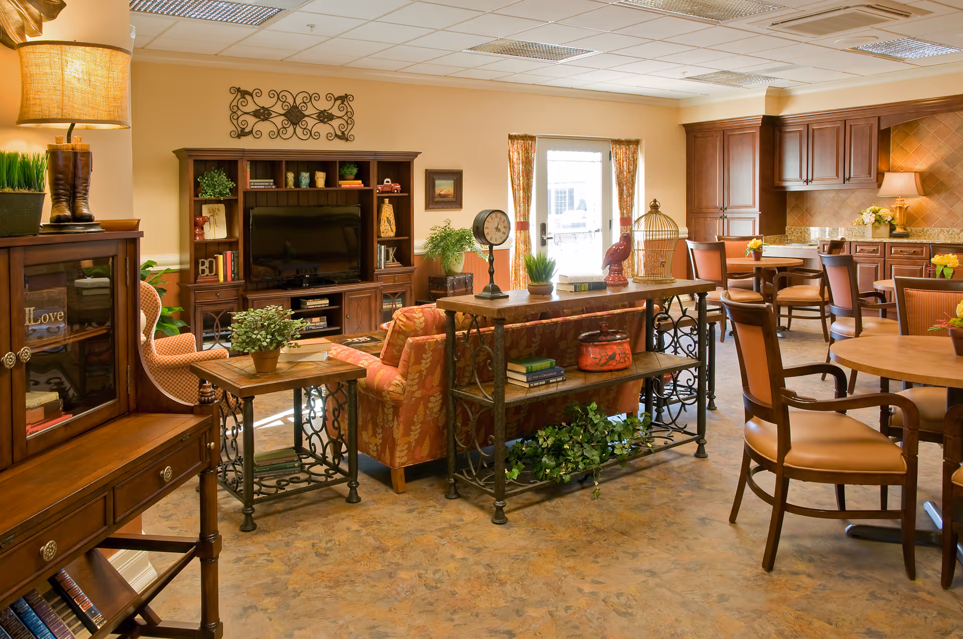 A cozy senior living common area featuring a seating arrangement with a floral-patterned sofa and armchair, wooden tables with decorative plants and books, a large wooden entertainment center with a flat-screen TV, and a kitchen area with wooden cabinets and dining tables with chairs. The room is warmly lit with natural light coming through a glass door with curtains.