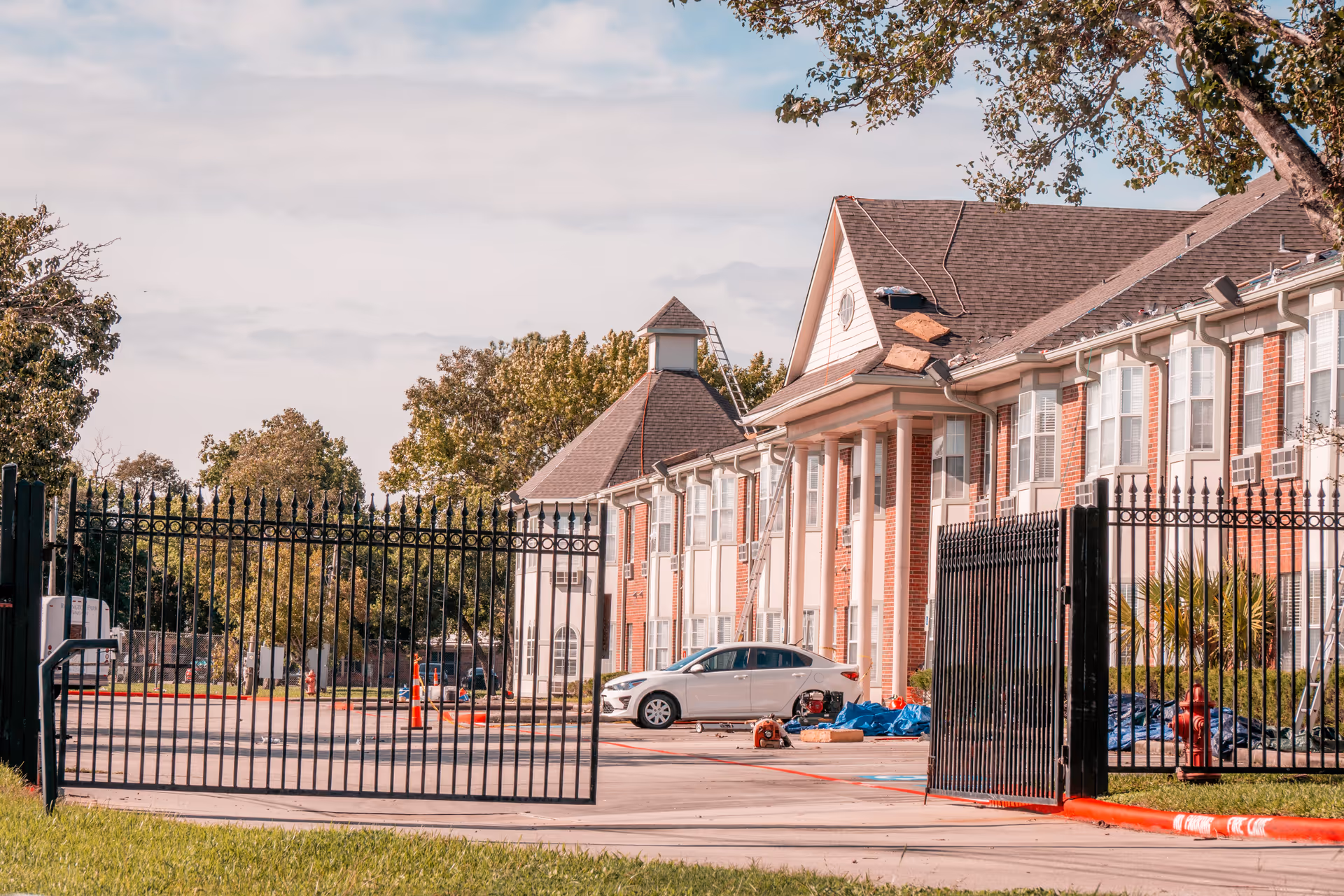 View of the exterior of a senior living facility named The Waterford at Baytown, showing a brick building with white columns and multiple windows. There is a black iron gate partially open in the foreground, a white car parked near the building, and some construction materials and ladders indicating roof repair work. Trees and a partly cloudy sky are visible in the background.