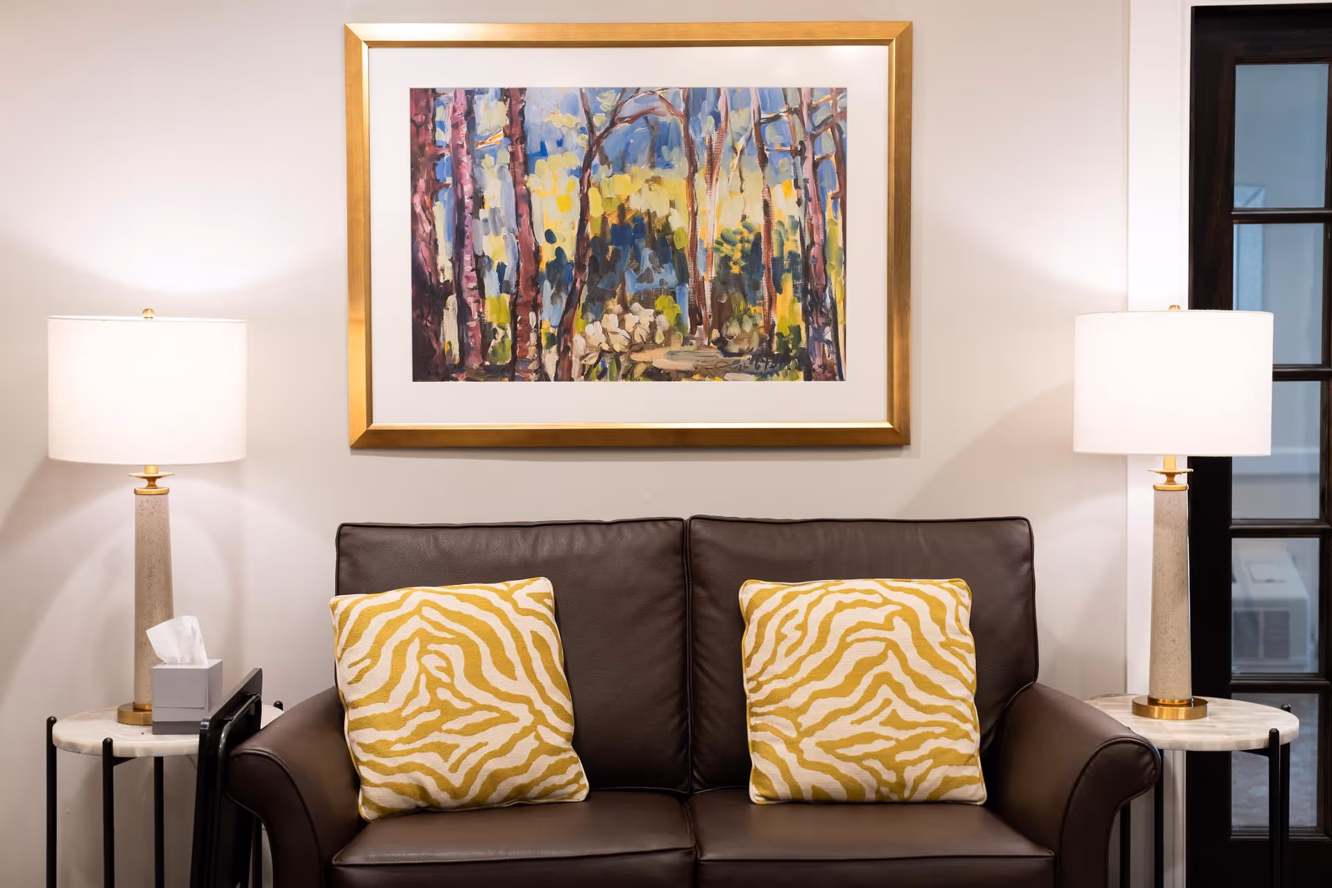 A brown leather loveseat with two yellow patterned pillows sits beneath a framed painting, flanked by matching table lamps on side tables.