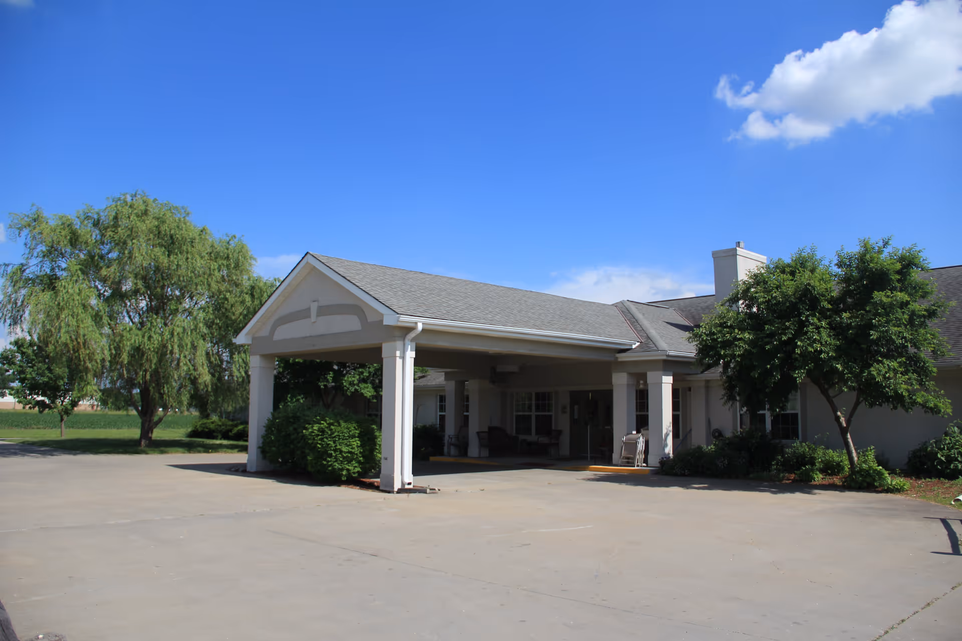 Exterior view of a single-story building with a covered entrance supported by white columns. The building is surrounded by green trees and bushes under a clear blue sky with a few clouds.