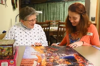 An elderly woman and a younger woman sitting at a table working together on a large jigsaw puzzle in a room with a dining area in the background.