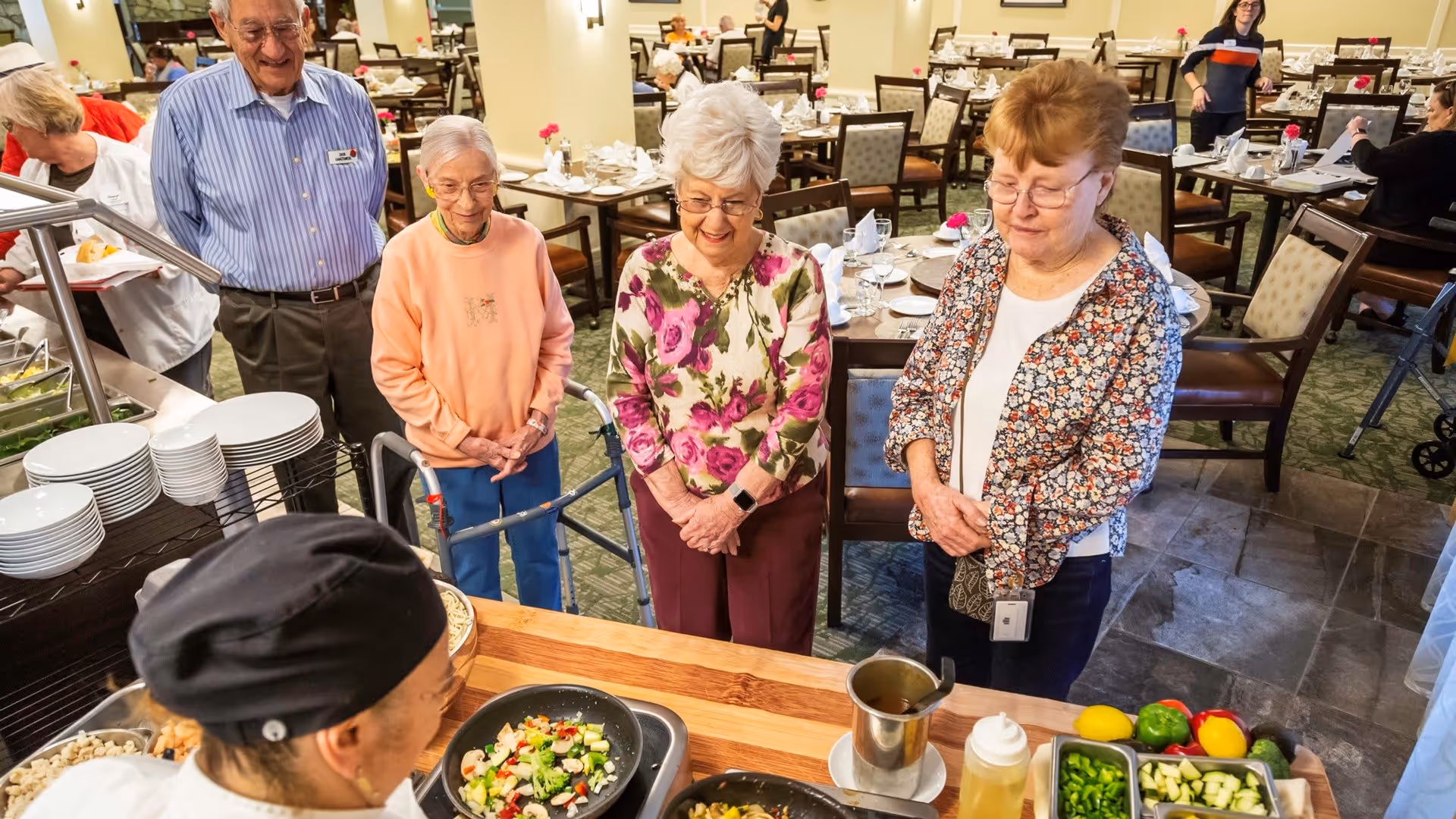 A group of elderly people standing at a food serving counter in a dining room, watching a chef prepare a meal with vegetables in a pan. The dining area is set with tables and chairs, and other people are visible in the background.