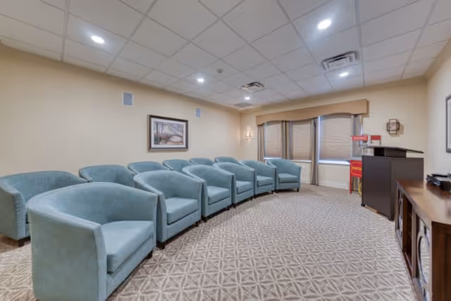 A small meeting or presentation room with two rows of light blue upholstered chairs arranged facing a wooden podium and a cabinet. The room has beige walls, a patterned carpet, recessed ceiling lights, a framed picture on the wall, and windows with blinds and a valance.