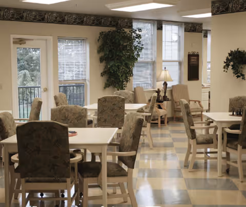 Communal dining room with multiple white tables and upholstered chairs, potted plants, windows, and soft lighting.