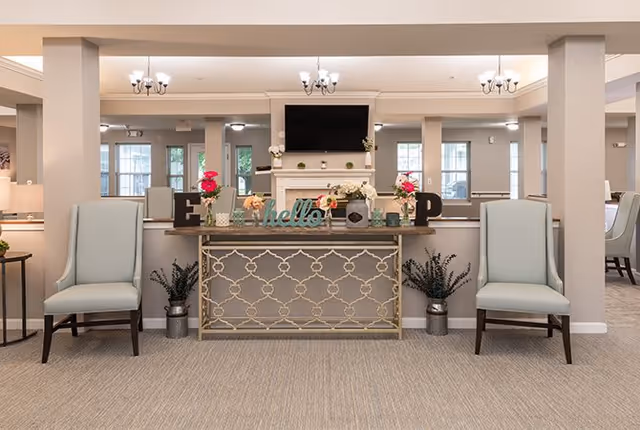 A welcoming interior space featuring a decorative console table with the word 'hello' and floral arrangements on top, flanked by two light blue upholstered chairs. In the background, there is a fireplace with a mounted flat-screen TV above it and multiple windows letting in natural light. The room has a neutral color palette with beige walls and carpeted flooring.