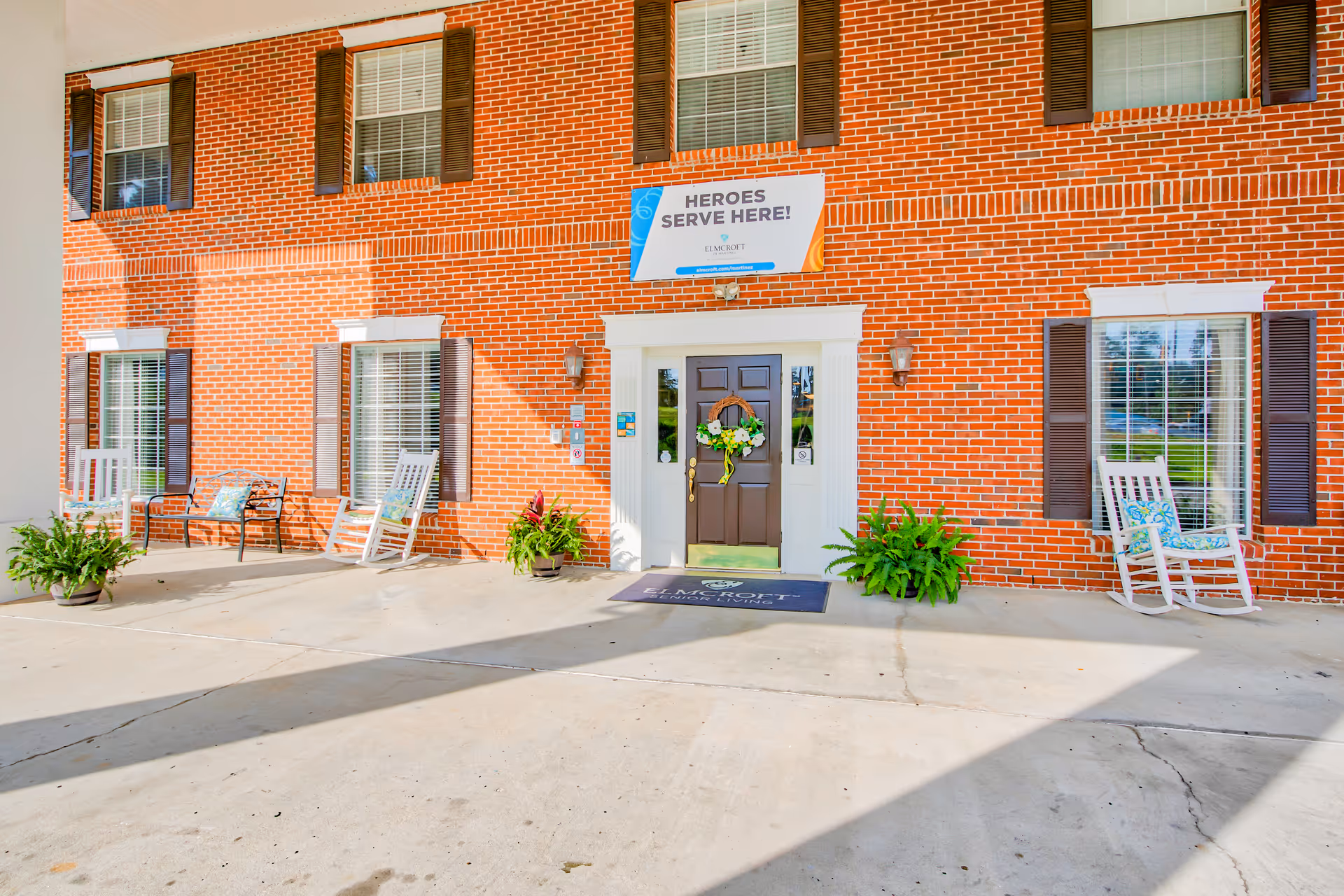 Front entrance of a red-brick senior living building with a wreath-adorned door, rocking chairs, potted plants, and a banner reading 'HEROES SERVE HERE!' above the entry.