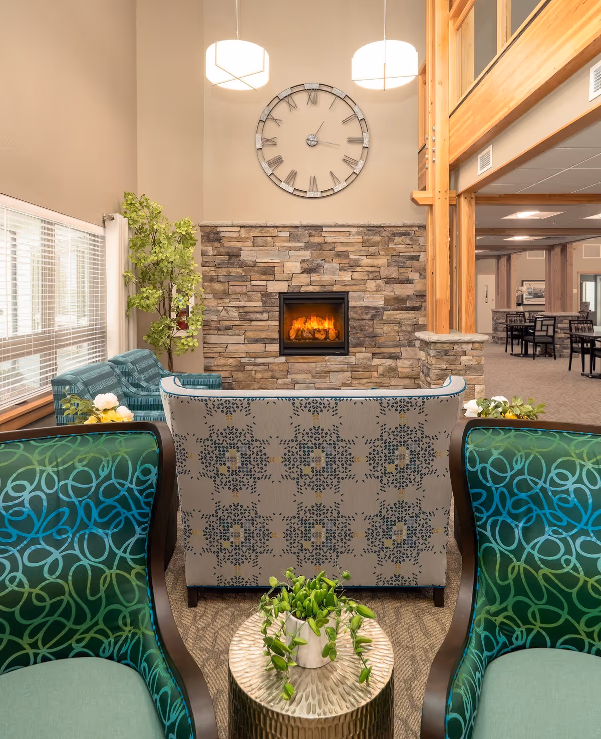 A cozy senior living community lounge area featuring a stone fireplace with a fire burning, a large wall clock above it, and comfortable seating including patterned armchairs and sofas. There is a small round table with a potted plant in the foreground, large windows with blinds on the left, and wooden beams and columns framing the space.