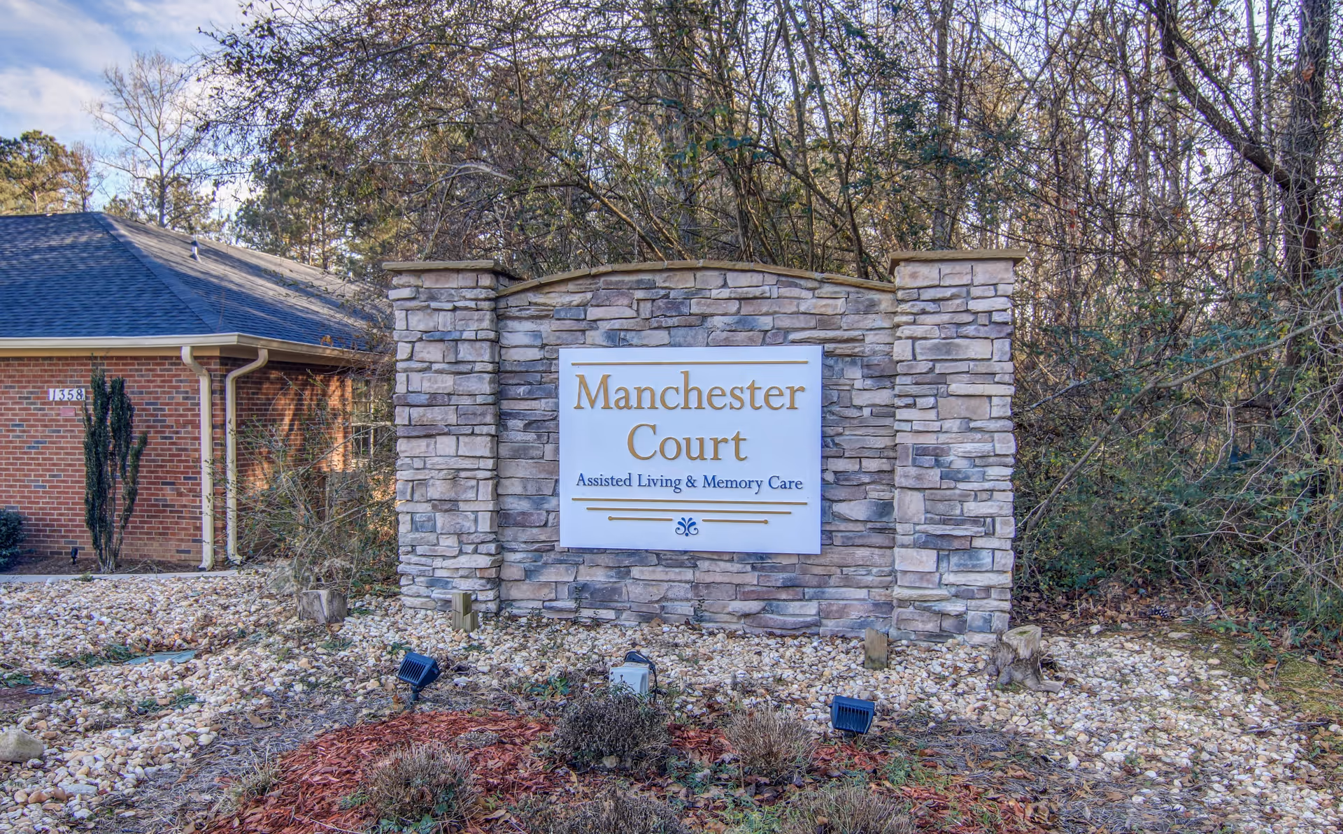 Stone sign for Manchester Court Assisted Living & Memory Care set in a landscaped area with rocks and plants, with trees and a brick building in the background.