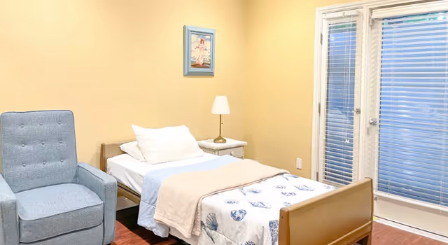 A small bedroom with a single bed covered with a seashell-patterned blanket and a beige throw. Next to the bed is a white nightstand with a lamp. A blue upholstered armchair is positioned to the left of the bed. On the wall above the nightstand is a framed picture. To the right, there are glass doors with white blinds.