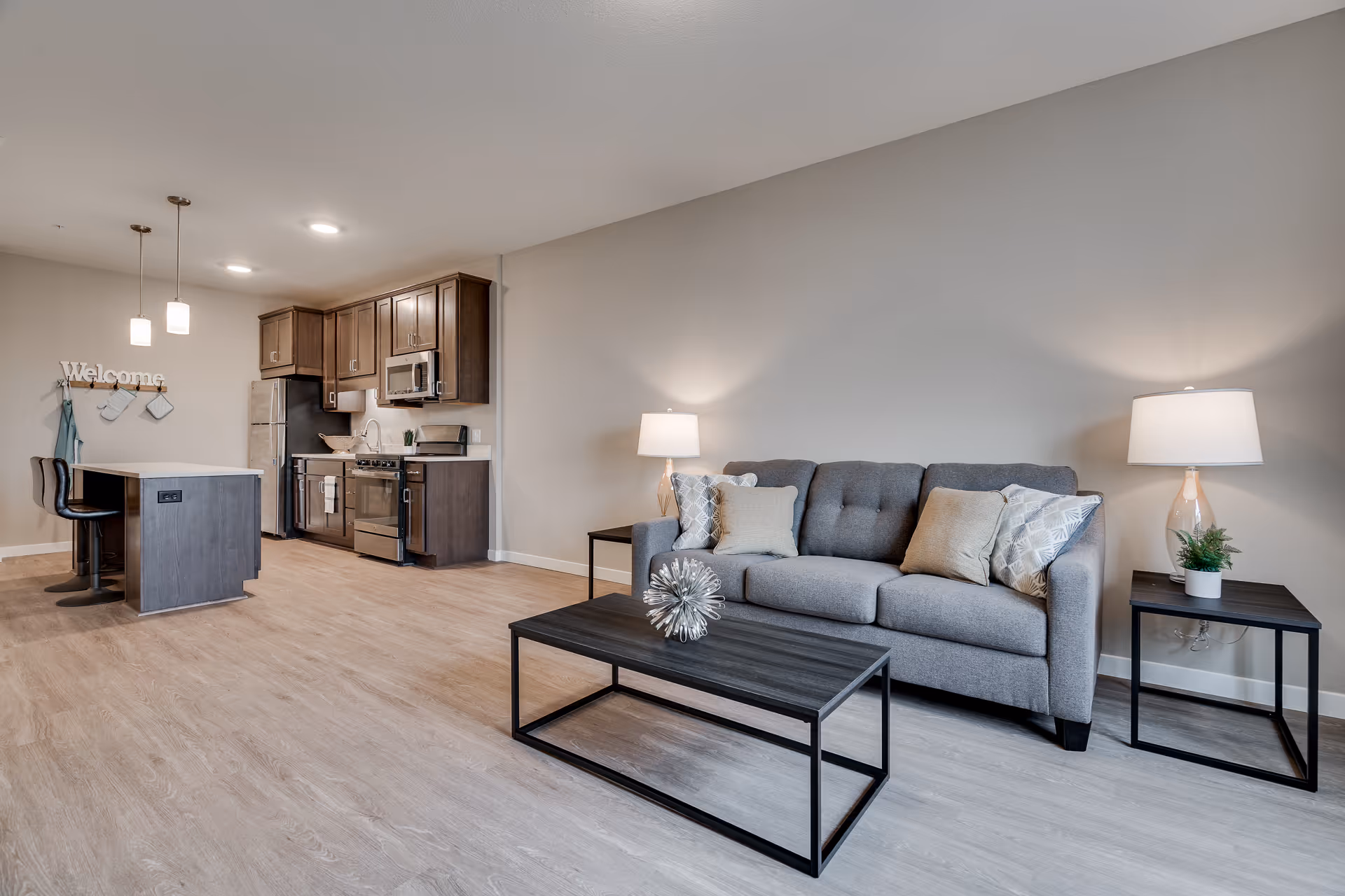 Open-plan living area with a gray sofa and coffee table in the foreground and a modern kitchen with an island and bar stools in the background.