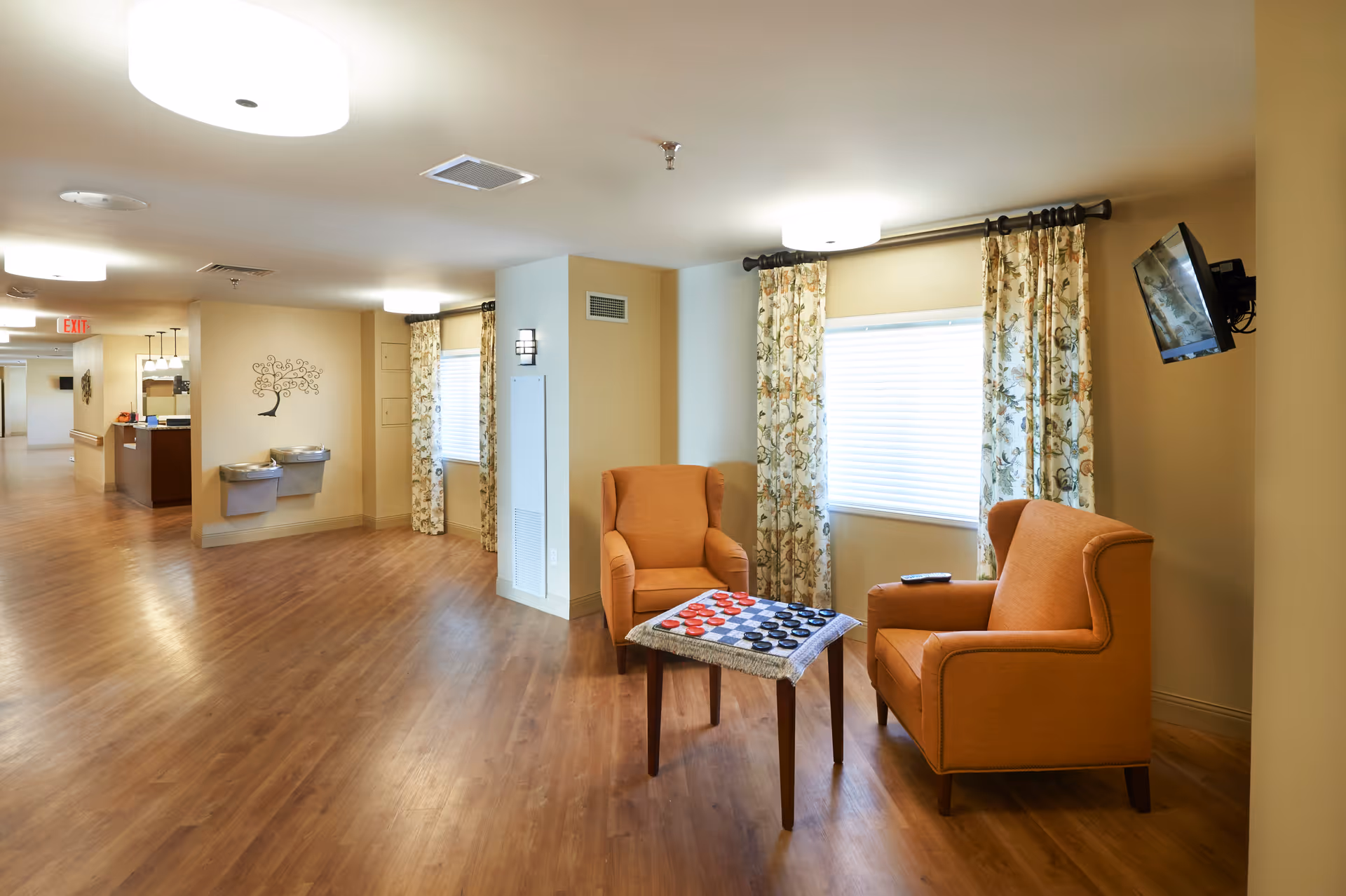A bright and spacious hallway area in a senior living facility with wooden flooring, two orange armchairs facing each other with a small table in between that has a checkers game set up. The walls are light-colored with floral curtains on the windows and a wall-mounted TV to the right. In the background, there are two water fountains and a decorative tree wall art, with a reception desk further down the hallway.