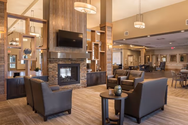 Spacious, warmly lit assisted living common area with seating arranged around a stone fireplace and wall-mounted TV.
