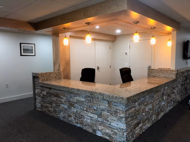 Reception area with a stone and granite counter, two black office chairs behind the counter, three pendant lights hanging from a wooden ceiling fixture, and two closed white doors in the background.