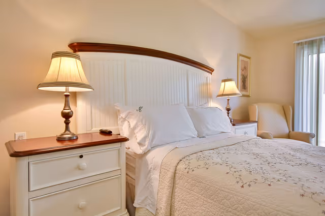 Bright bedroom featuring a neatly made bed with a white headboard, two nightstands with lamps, and an armchair by sliding glass doors.