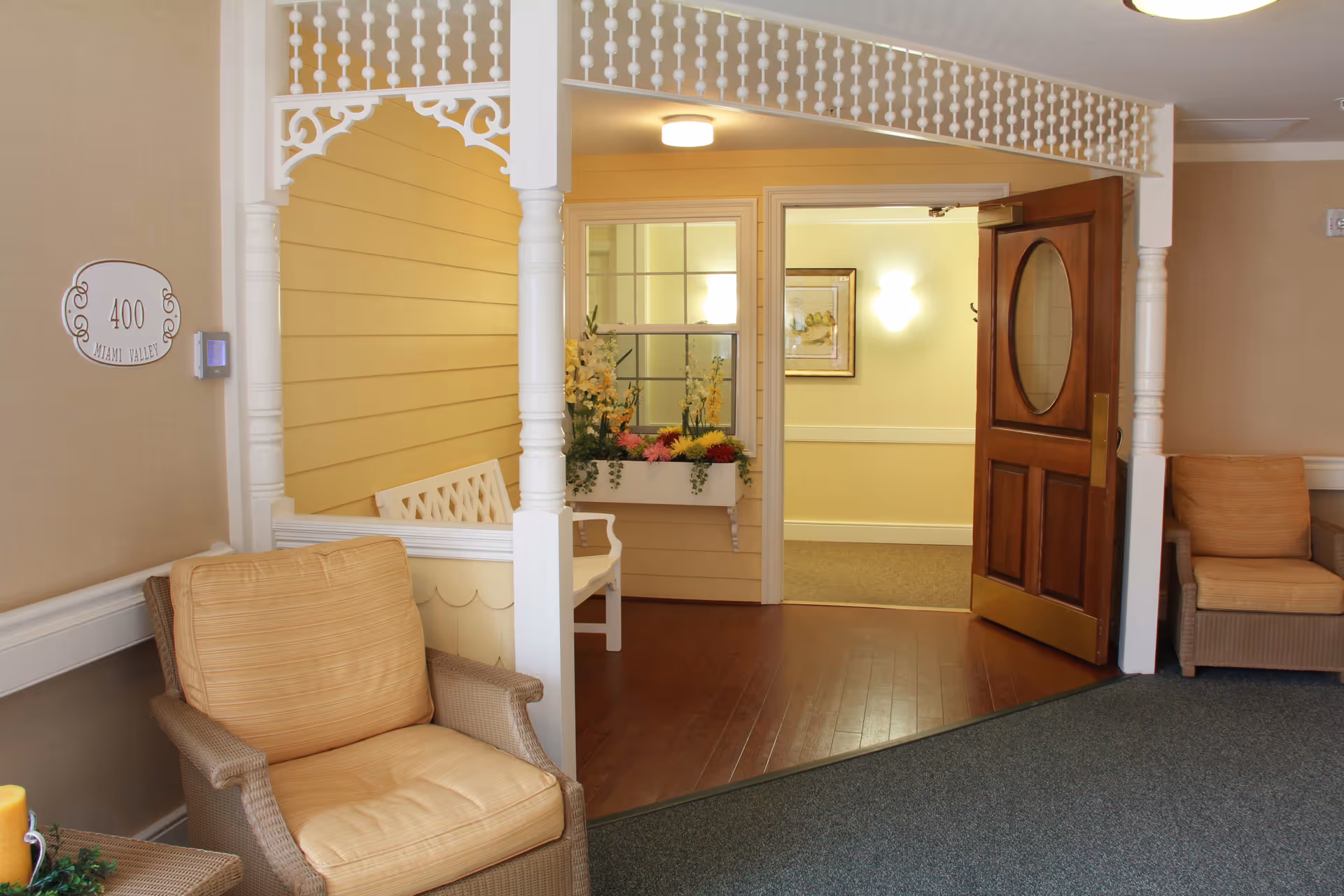 A cozy interior seating area in a senior living facility with two cushioned armchairs, a decorative white wooden bench, and a wooden door with an oval window leading to a hallway. The walls are painted in warm tones with floral decorations and framed artwork visible in the hallway.