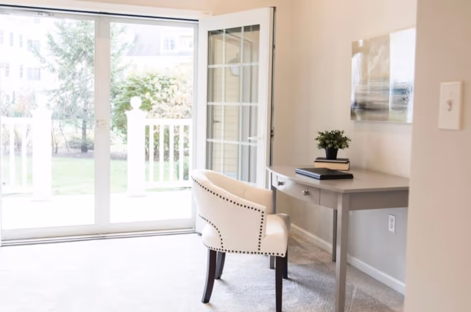 A bright interior room with a light gray desk and a cream upholstered chair with nailhead trim. On the desk are two closed books and a small potted plant. A large glass sliding door and a glass door with white frames open to a green outdoor area with trees and bushes. A modern abstract painting hangs on the beige wall above the desk.