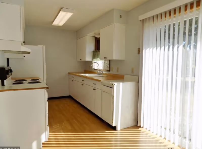 Sunlit kitchen with white cabinets, a stove and sink, and vertical sliding blinds over a glass door.
