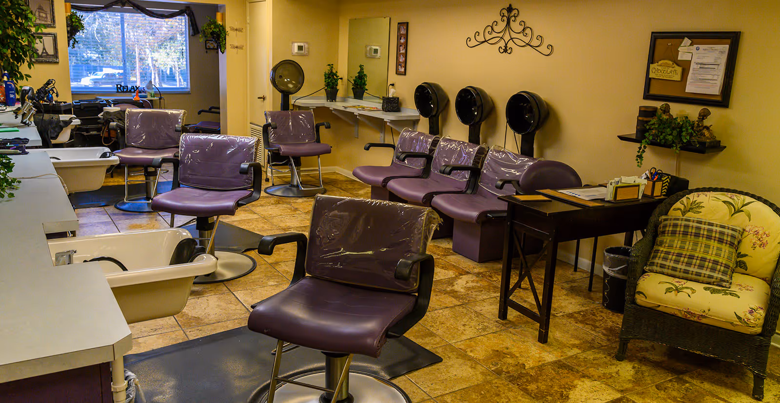 Interior view of a hair salon area with multiple purple salon chairs, hair washing sinks, and hair dryers mounted on the wall. There is a window with a 'RELAX' sign, a small desk with office supplies, and a cushioned chair with a plaid pillow. The floor is tiled and the walls are painted yellow.