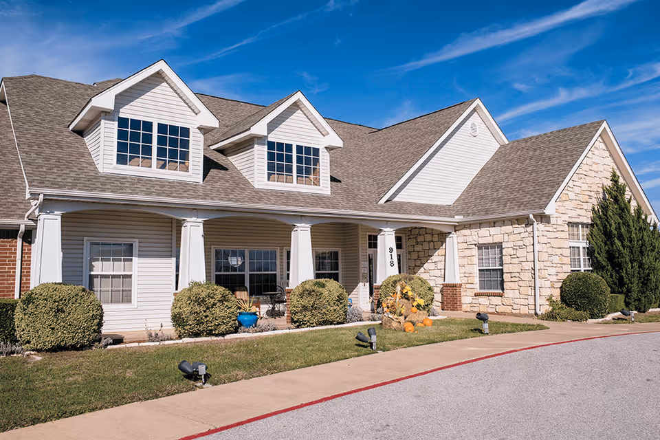 Exterior view of a single-story senior living facility building with a combination of stone and siding walls, multiple windows, a covered porch with white columns, neatly trimmed bushes, and a clear blue sky.