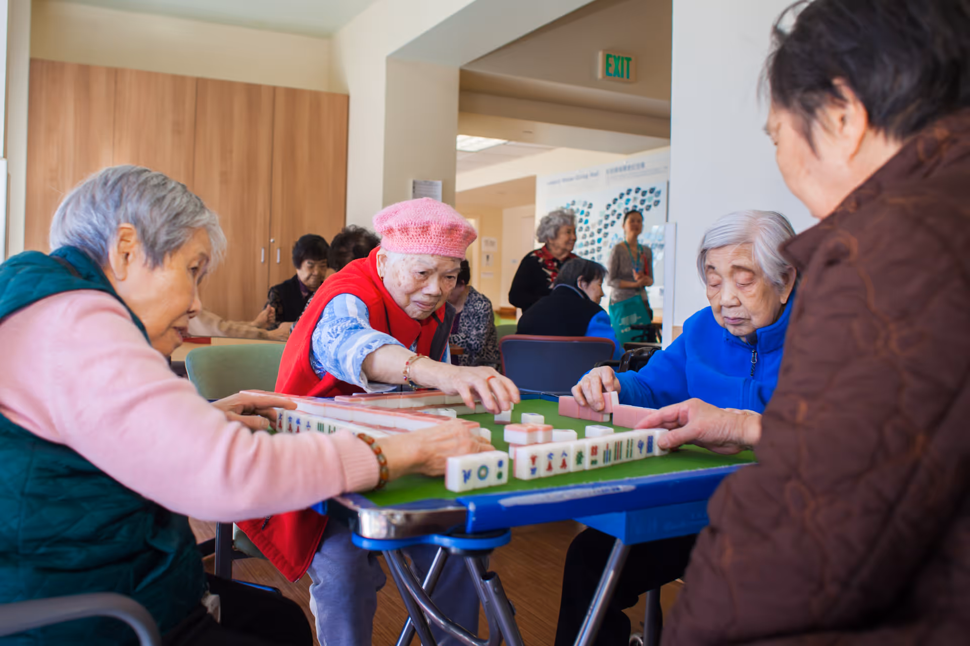 A group of elderly women sitting around a table playing a game of Mahjong in a well-lit room with wooden flooring and light-colored walls. Some other elderly people and a staff member are visible in the background.