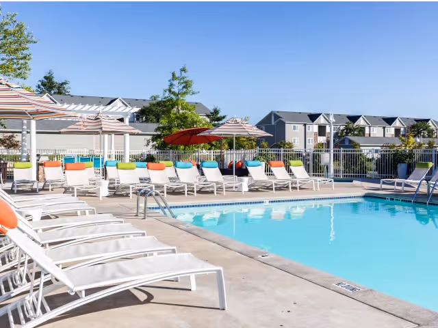 Outdoor swimming pool area with multiple white lounge chairs lined up along the poolside, some with colorful cushions. Several large umbrellas provide shade, and residential buildings are visible in the background under a clear blue sky.