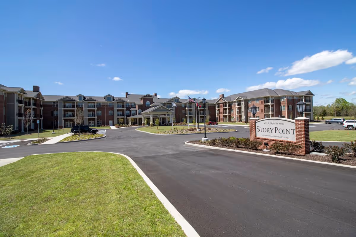 Exterior view of StoryPoint Collierville senior living facility showing a large three-story brick building with balconies, a circular driveway, landscaped grass and shrubs, and a clear blue sky.