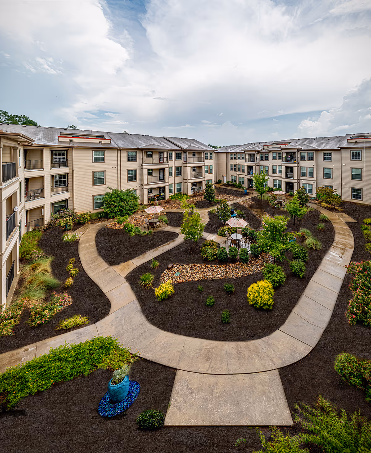 View of an outdoor courtyard area at Parkview on Hollybrook featuring winding concrete pathways, landscaped garden beds with various shrubs and small trees, several patio tables with umbrellas, and a three-story residential building surrounding the courtyard under a partly cloudy sky.