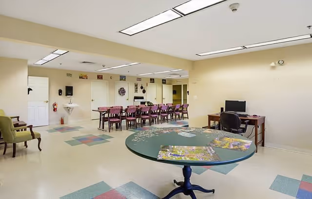 A spacious room with beige walls and a patterned floor featuring pastel-colored tiles. In the foreground, there is a round green table with puzzles on it. In the background, there is a long table with multiple chairs arranged around it. A computer desk with a chair and monitor is positioned against the right wall. On the left side, there are two armchairs and a wall-mounted sink with a soap dispenser and fire extinguisher nearby. Several doors and wall decorations are visible along the back wall.