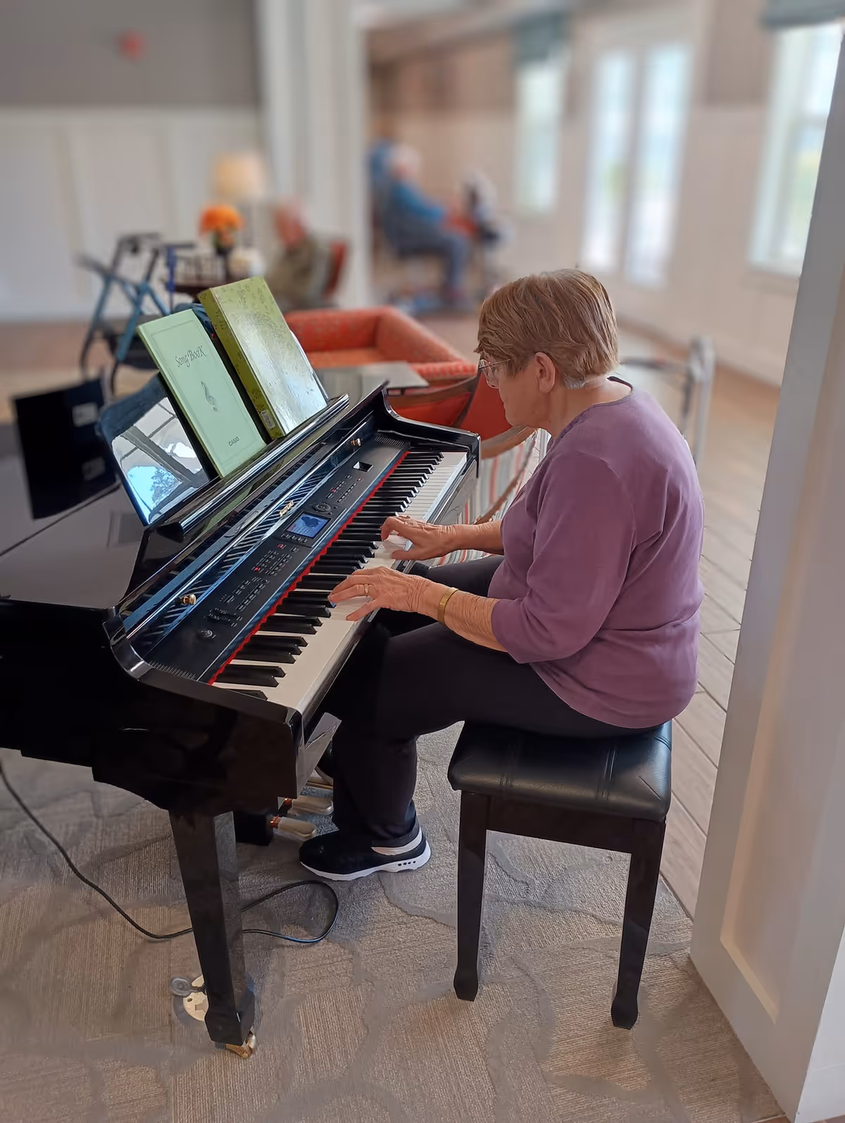 An elderly woman wearing a purple top and black pants is seated on a black piano bench playing a black grand piano with sheet music on the stand. The room has large windows letting in natural light, and there are other elderly people seated in the background on chairs and sofas.