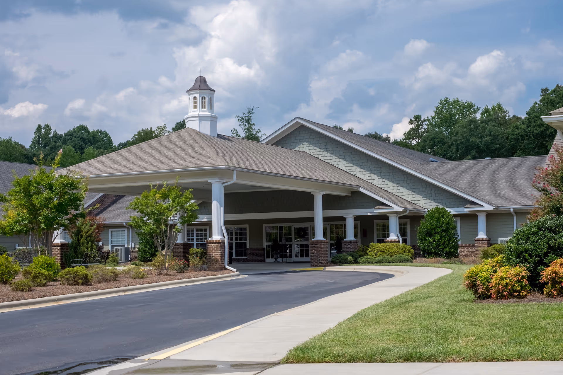 Front exterior view of a senior living facility named White Oak of Waxhaw, featuring a covered entrance with white columns, brick accents, landscaped greenery, and a driveway leading up to the entrance under a partly cloudy sky.
