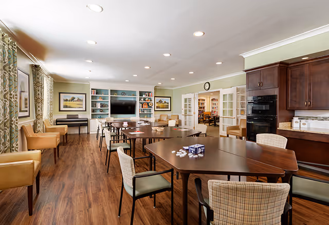 A spacious senior living common area with multiple tables and chairs arranged for group activities. The room features wood flooring, light green walls, and built-in shelves with a large flat-screen TV. There are comfortable armchairs along the walls, framed artwork, and a kitchen area with dark wood cabinets and appliances. French doors lead to another room with bookshelves.