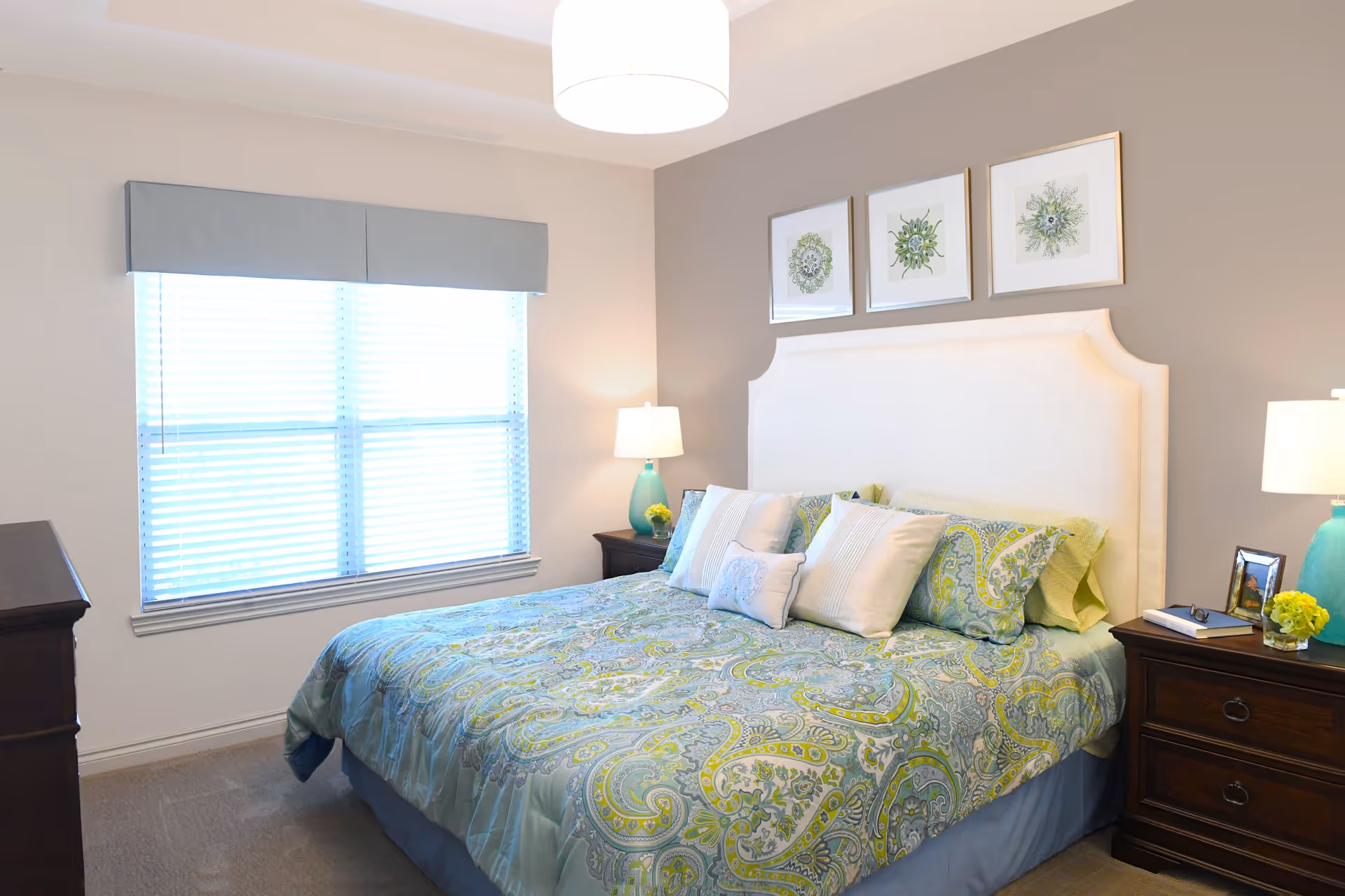 A bright bedroom with a large bed featuring a paisley-patterned bedspread in shades of blue and green. The bed has a white upholstered headboard and is flanked by two dark wooden nightstands, each with a turquoise lamp and decorative items. Above the headboard are three framed botanical prints. A window with white blinds and a light blue valance is on the left wall.
