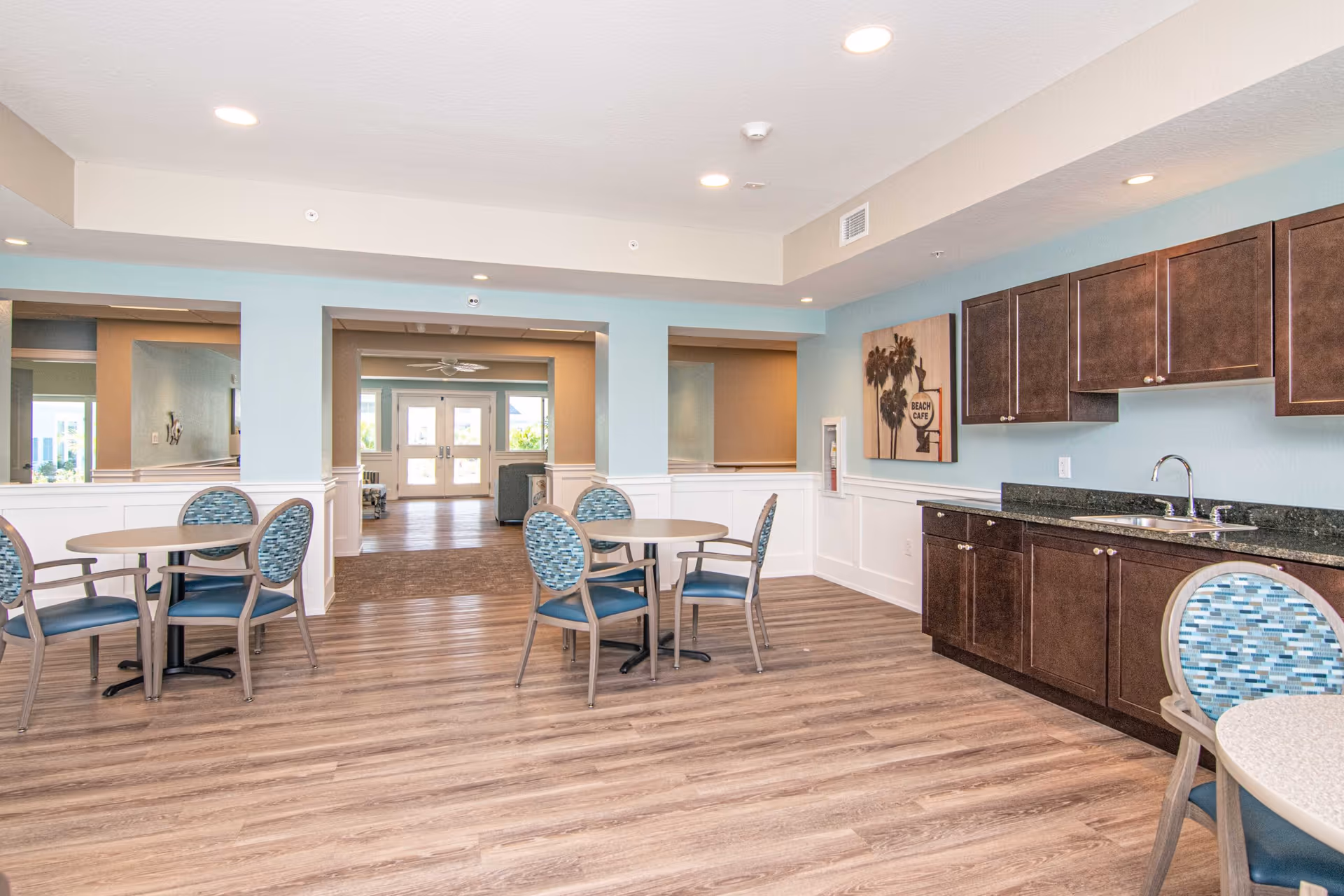 Interior view of a senior living facility common area with light blue walls, wood flooring, round tables with blue cushioned chairs, and a kitchenette with dark wood cabinets and a sink. There is a painting on the wall and a hallway leading to double doors with windows.
