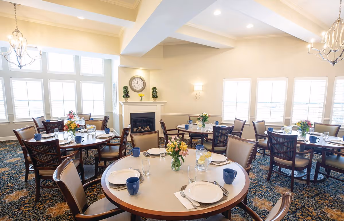 Bright communal dining room with round tables set for meals, floral centerpieces, chairs, and a fireplace beneath large windows.