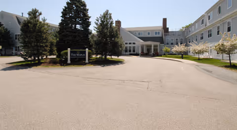 Exterior view of a senior living facility named The Maples, featuring a large paved driveway, several tall evergreen trees, and a multi-story building with white siding and multiple windows under a clear blue sky.
