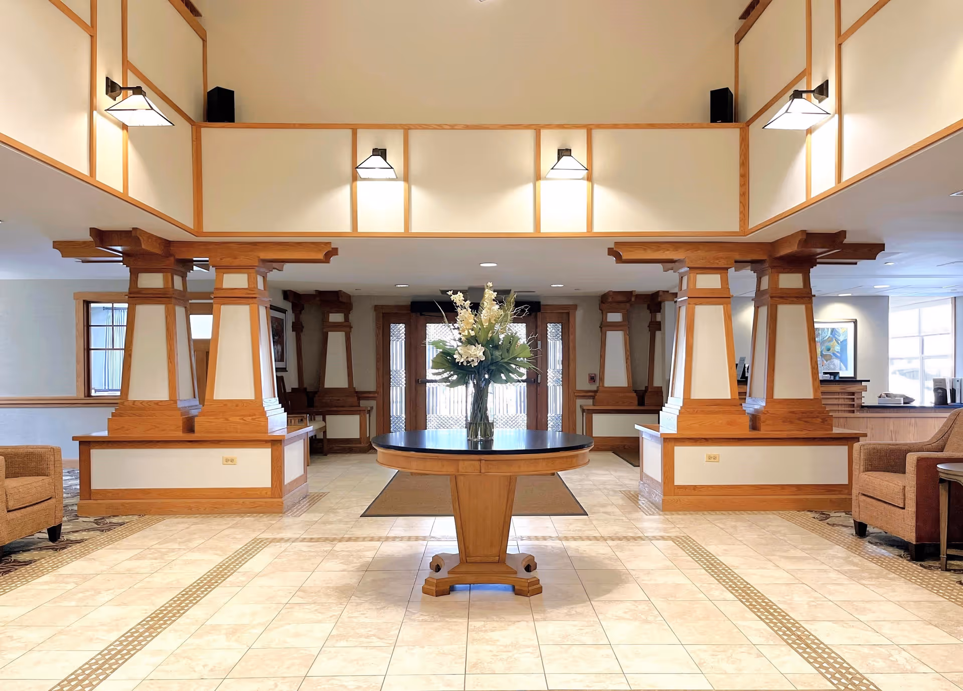 Spacious and well-lit lobby area with a round wooden table in the center holding a vase of white flowers. The room features beige tiled flooring with decorative patterns, wooden pillars with white panels, wall-mounted lights, and comfortable seating areas with armchairs on either side. Large glass doors are visible at the far end of the lobby.
