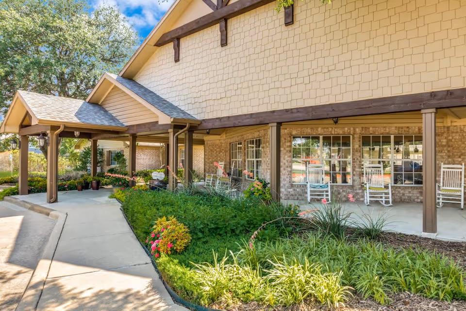 Exterior view of a senior living facility with a covered walkway, rocking chairs on the porch, and landscaped garden beds with green plants and flowers under a clear blue sky.
