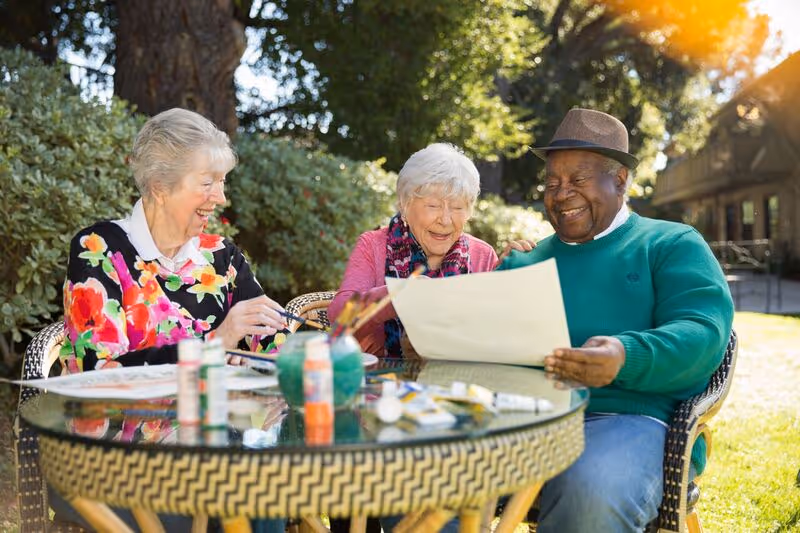 Three elderly people sitting outdoors around a glass-top table with art supplies, smiling and looking at a large sheet of paper. They are surrounded by greenery and sunlight.